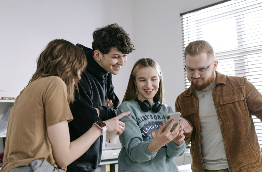 Four young adults smiling and gathered around a phone, reading something together.