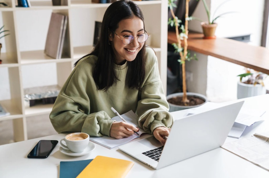 A smiling person in a green sweater working at a laptop and taking notes at a bright desk.